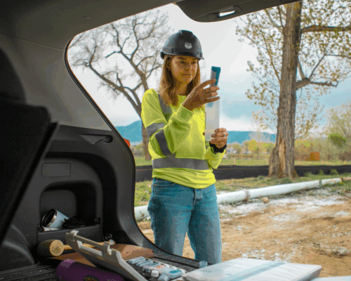Woman in safety gear examines sample near construction materials outdoors.