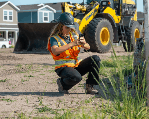 Worker in hard hat takes photo at construction site; loader, houses behind.