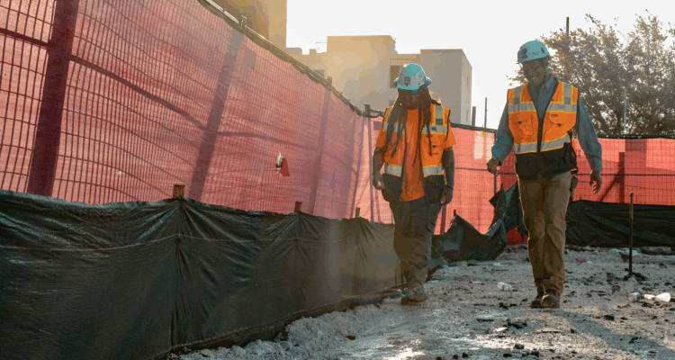 Two workers in orange vests walk along a fenced construction site.
