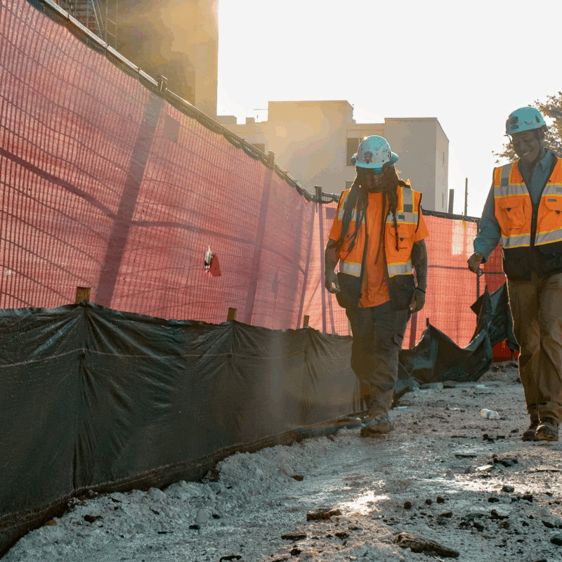 Two workers in orange vests walk along a fenced construction site.