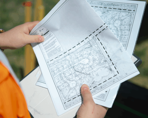 Person in orange jacket reviews construction project plans outdoors.