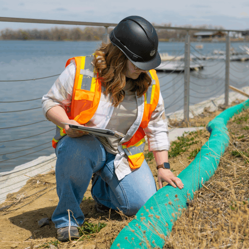 Person in hard hat inspects green barrier by riverbank.