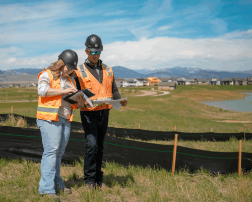 Two construction workers in safety vests review documents at outdoor site.