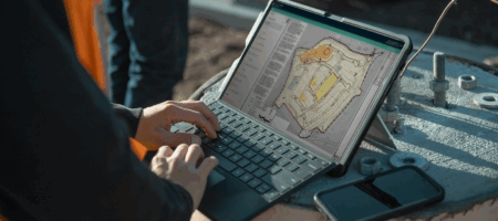 Person outdoors uses laptop, project plans on screen, phone nearby.