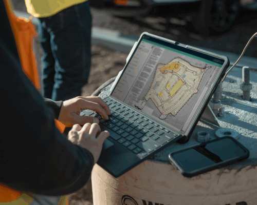 Person outdoors uses laptop, project plans on screen, phone nearby.