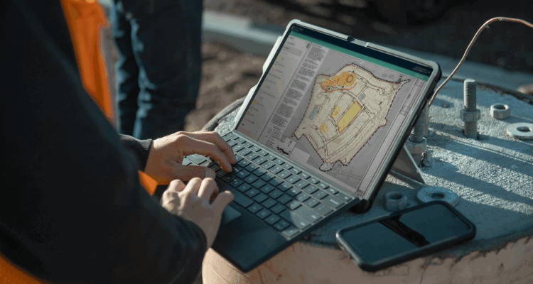 Person outdoors uses laptop, project plans on screen, phone nearby.