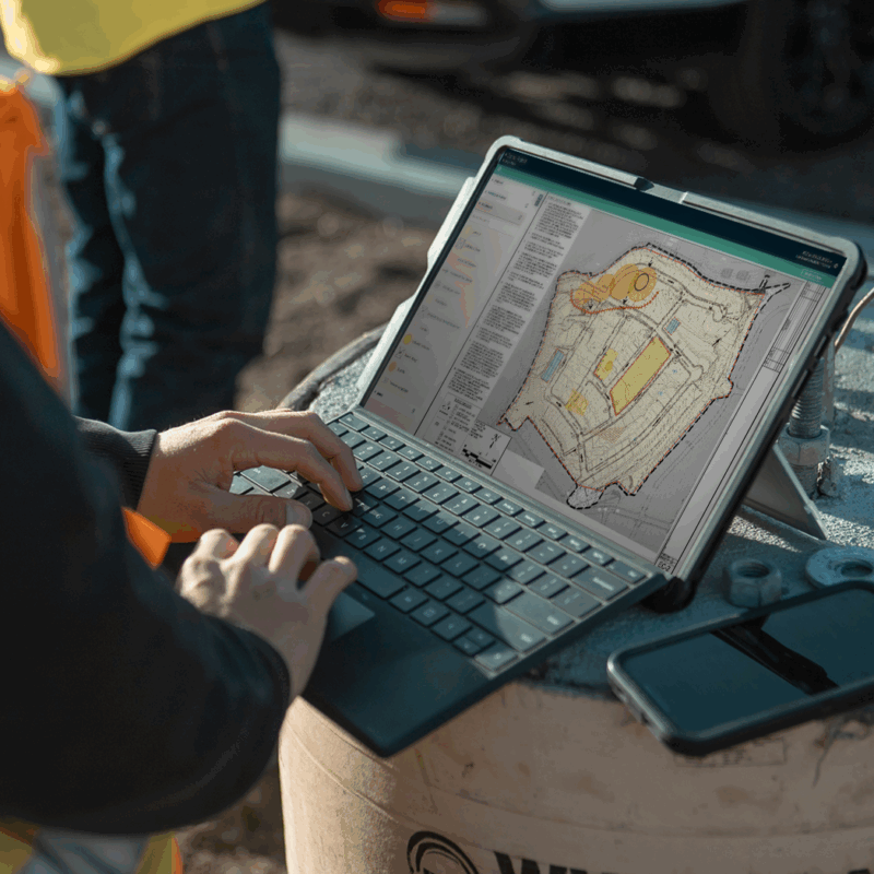 Person outdoors uses laptop, project plans on screen, phone nearby.