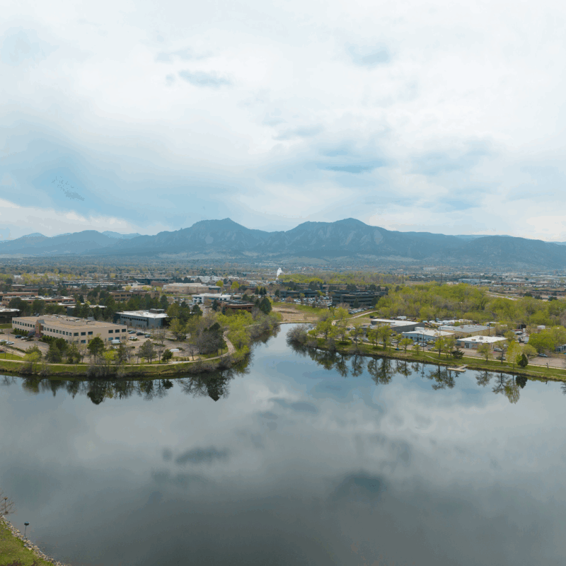 Lake reflects trees, buildings, clouds, and distant mountains.
