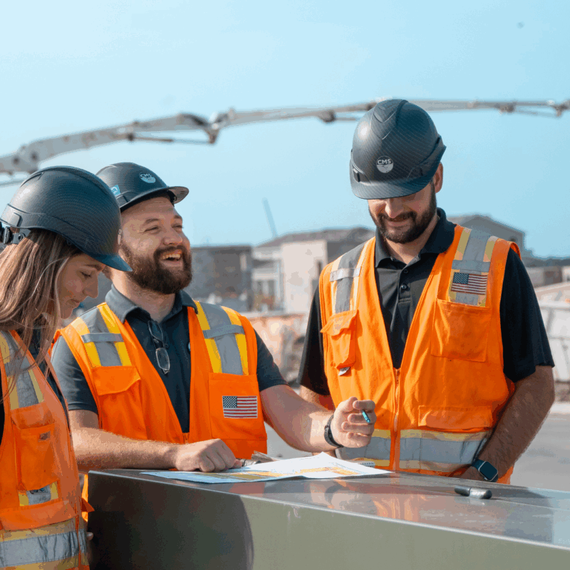 Three workers in orange vests review plans at construction site.