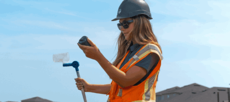 Woman in safety vest examines container by water.