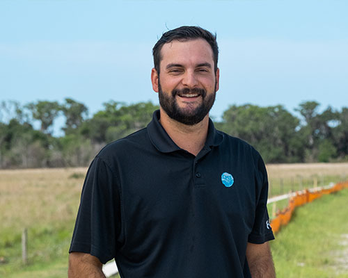 Bearded man in black polo smiles outside by grass and trees.