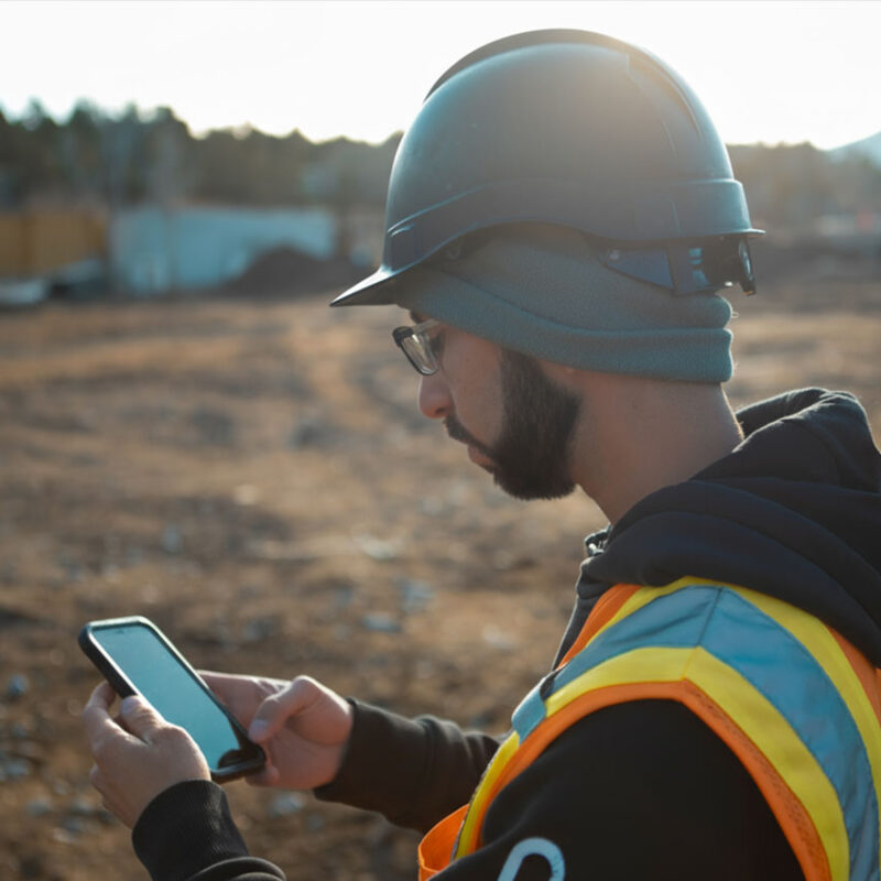 Person in hard hat and vest uses smartphone at construction site.
