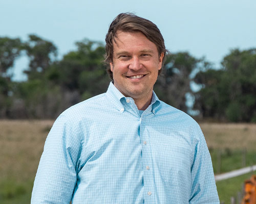 Man in light blue checkered shirt smiles outdoors by trees.