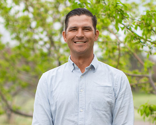 Man in blue shirt smiles outdoors before green trees.
