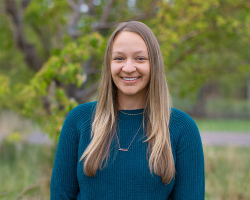 Smiling woman in blue sweater outdoors with greenery.