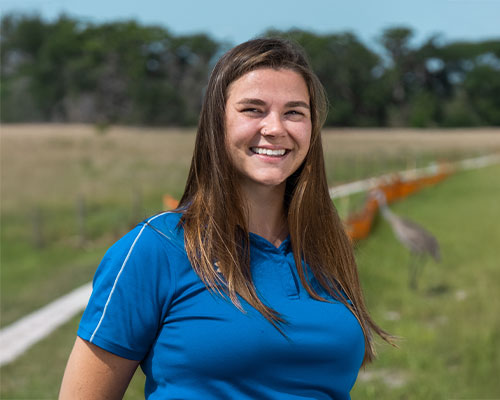 Woman in blue polo stands on grassy field with trees.