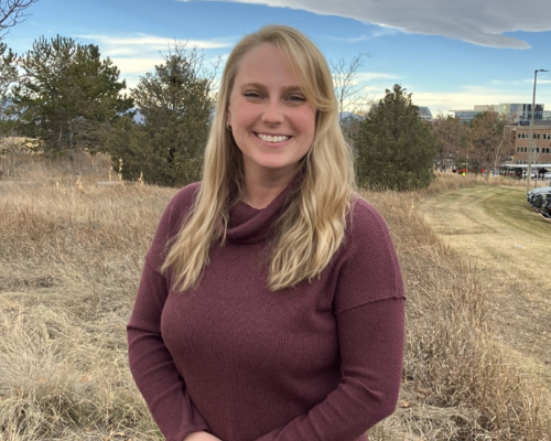 Blonde woman in maroon sweater stands outside on grass.