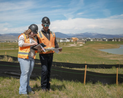 Two construction workers in safety vests review documents at outdoor site.