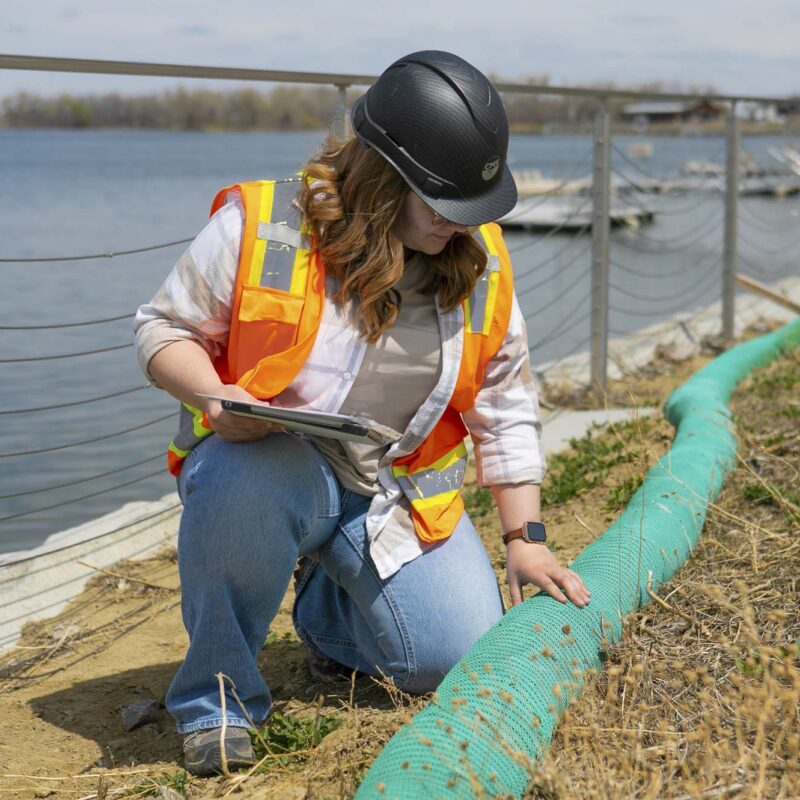Person in hard hat inspects green barrier by riverbank.
