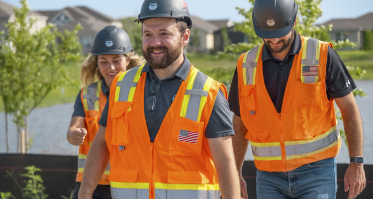 Three people in orange safety vests walk at construction site.