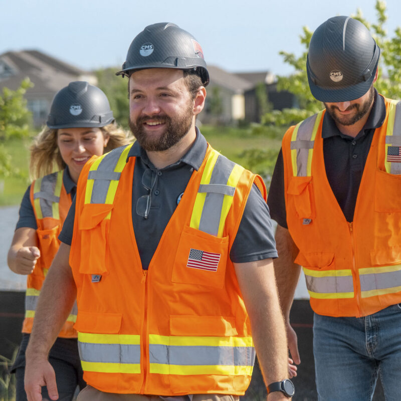 Three people in orange safety vests walk at construction site.