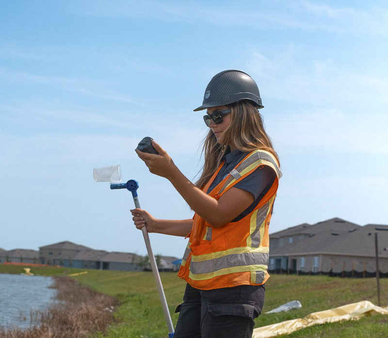 Woman in safety vest examines container by water.